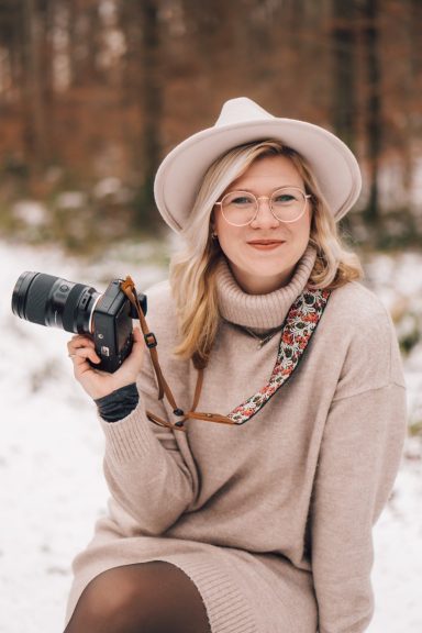 Frau mit Kamera in einem winterlichen Wald, trägt einen weißen Hut und einen beigen Pullover.
