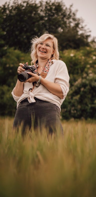 Lächelnde Frau mit Kamera in einem Feld mit hohen Gräsern.
