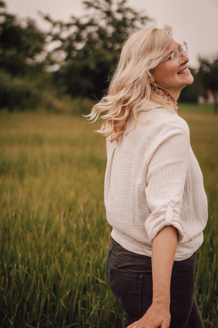 Lächelnde Frau mit blonden Haaren in einem Feld, schaut über die Schulter.